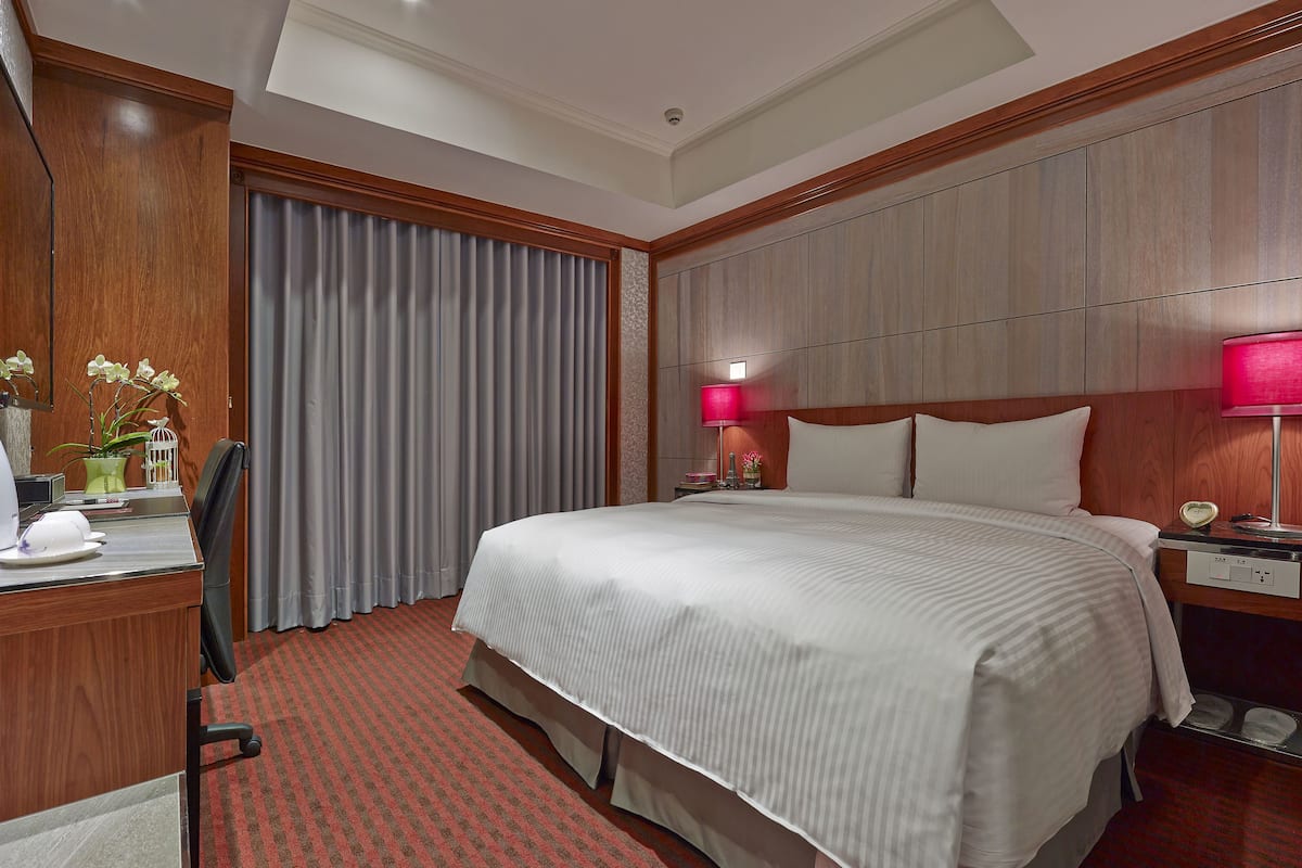 Ein Hotelzimmer mit einem großen Bett, einem Schreibtisch mit Stuhl, einem Nachttisch mit Lampe und einem Fenster mit Vorhängen.