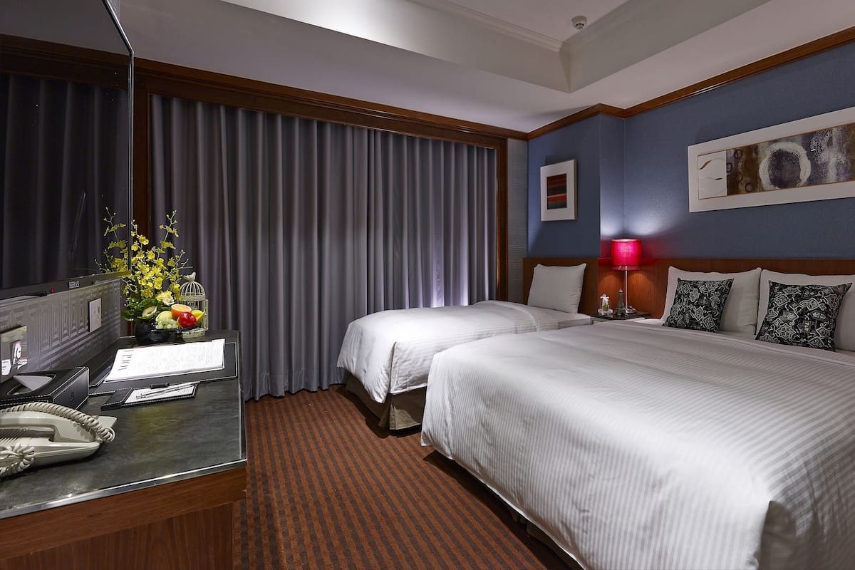 Ein Hotelzimmer mit zwei Betten, einem Schreibtisch mit Telefon, einer Lampe und einer Vase mit Blumen.