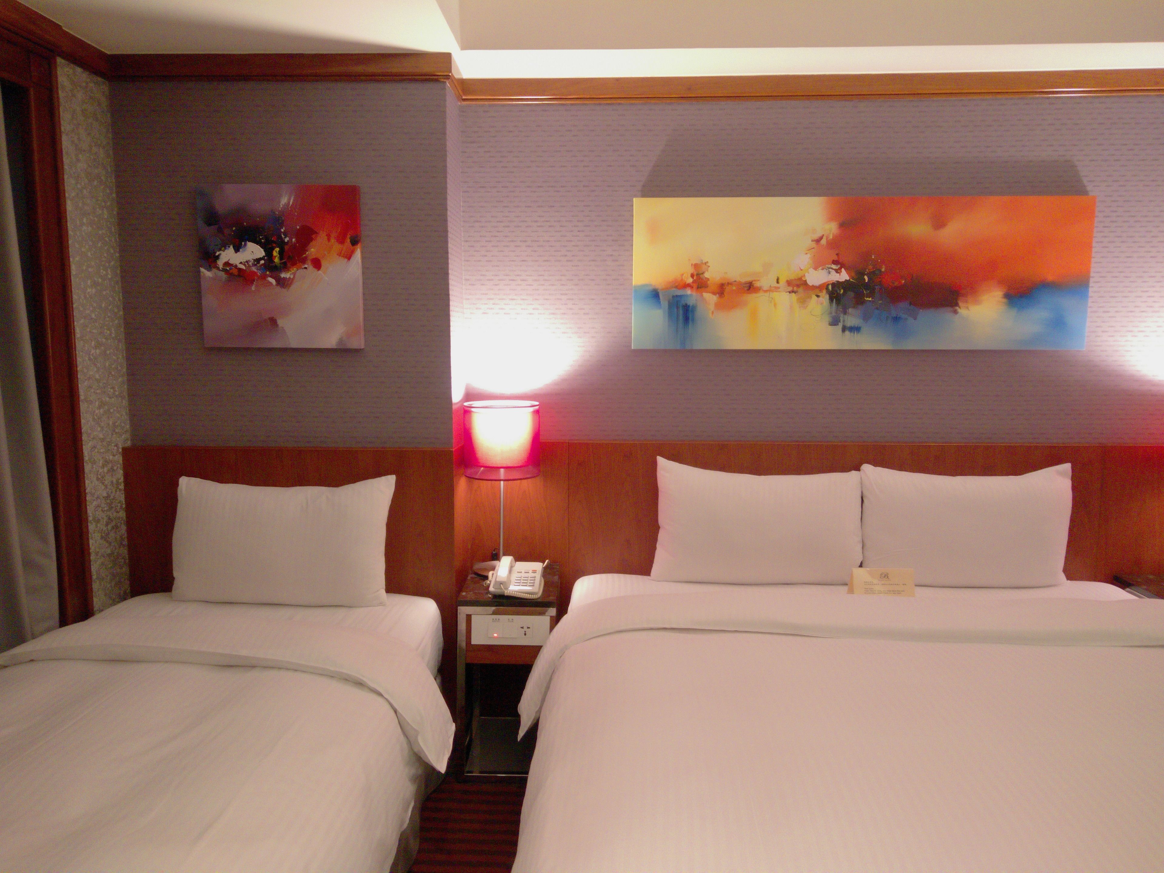 A hotel room with two beds, a nightstand with a lamp, and two paintings on the wall.