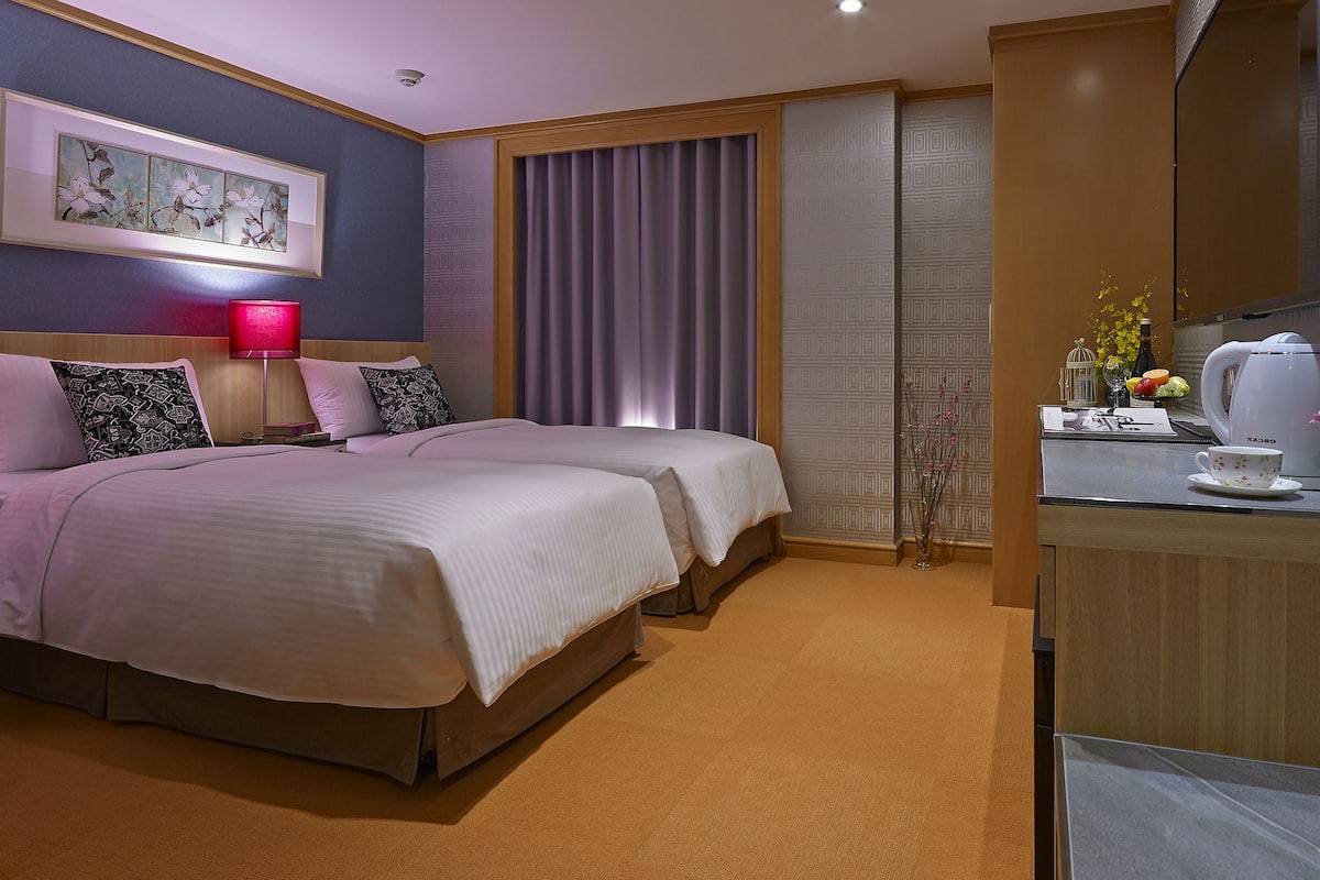 Ein Hotelzimmer mit zwei Betten, einer roten Lampe, einem gerahmten Kunstwerk, einem Fenster mit Vorhängen, einem kleinen Tisch mit Wasserkocher und einer Vase mit Blumen.