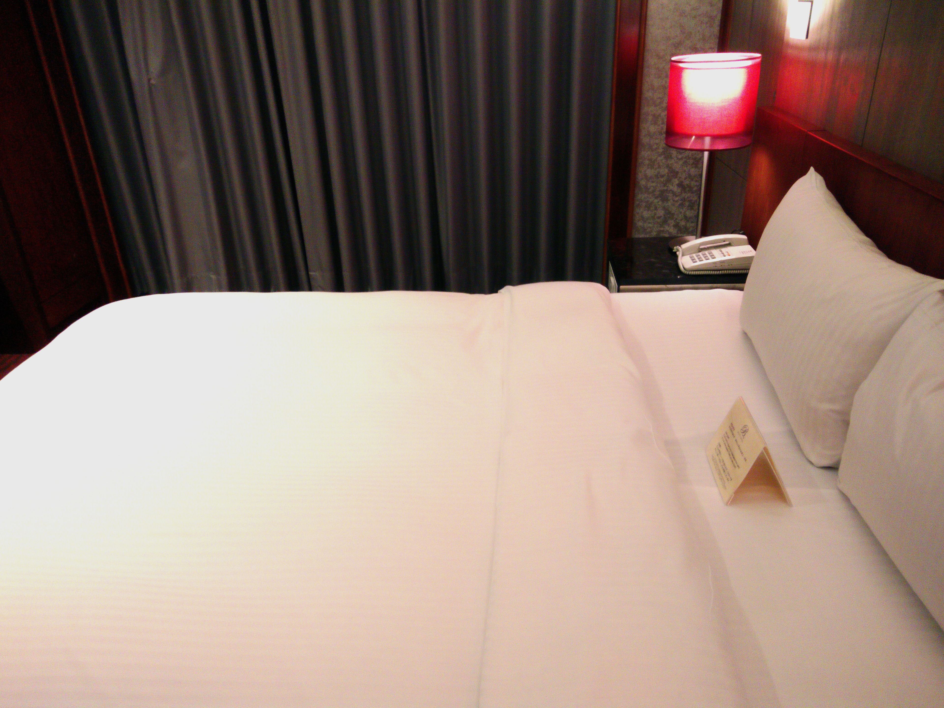 A hotel room with a neatly made bed, a bedside lamp, and a telephone on the nightstand.