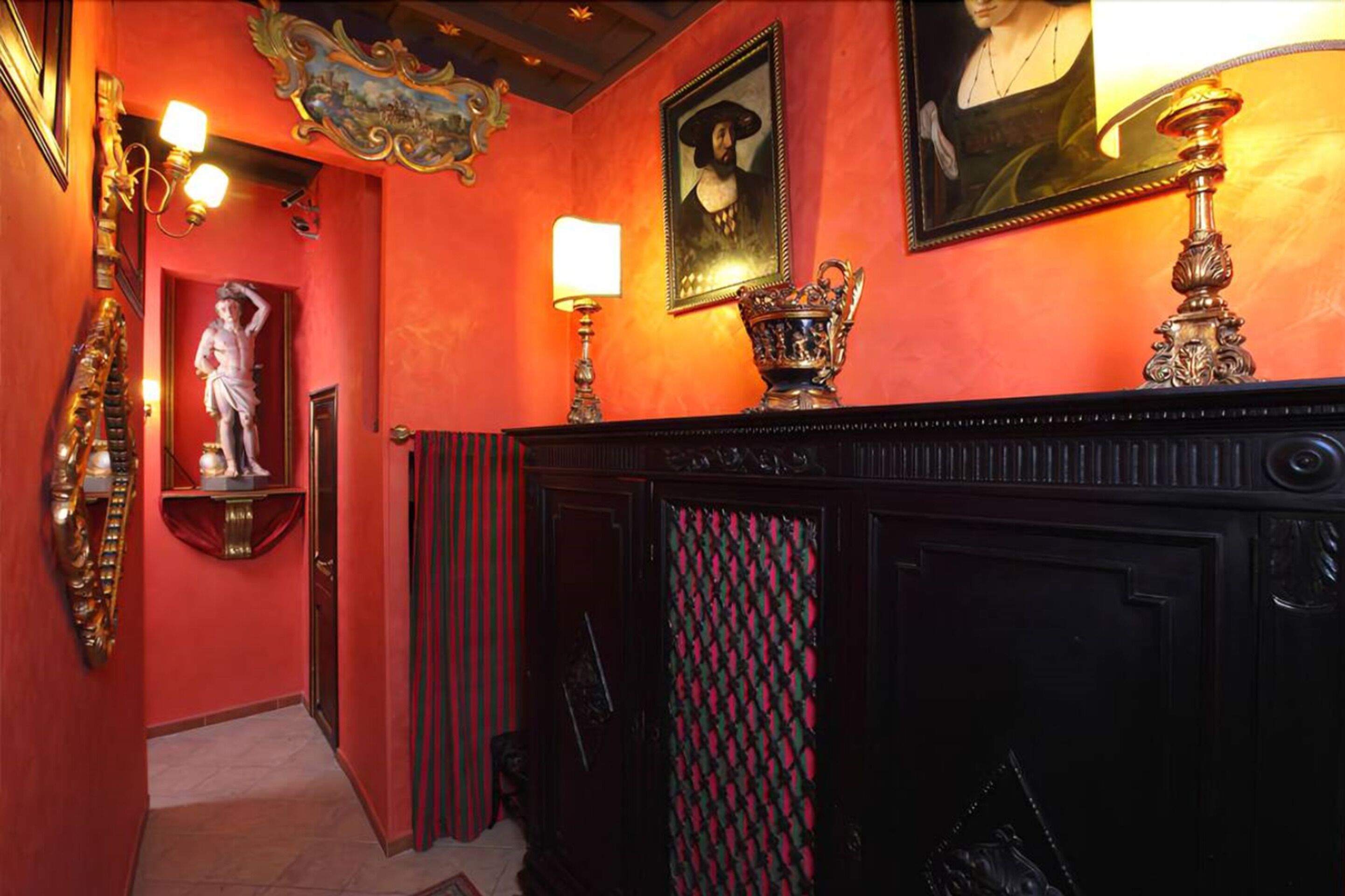 Un couloir avec un mur rouge, une statue, une cheminée ornée d’un rideau à motifs et un portrait encadré.