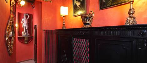 Un couloir avec un mur rouge, une statue, une cheminée ornée d’un rideau à motifs et un portrait encadré.