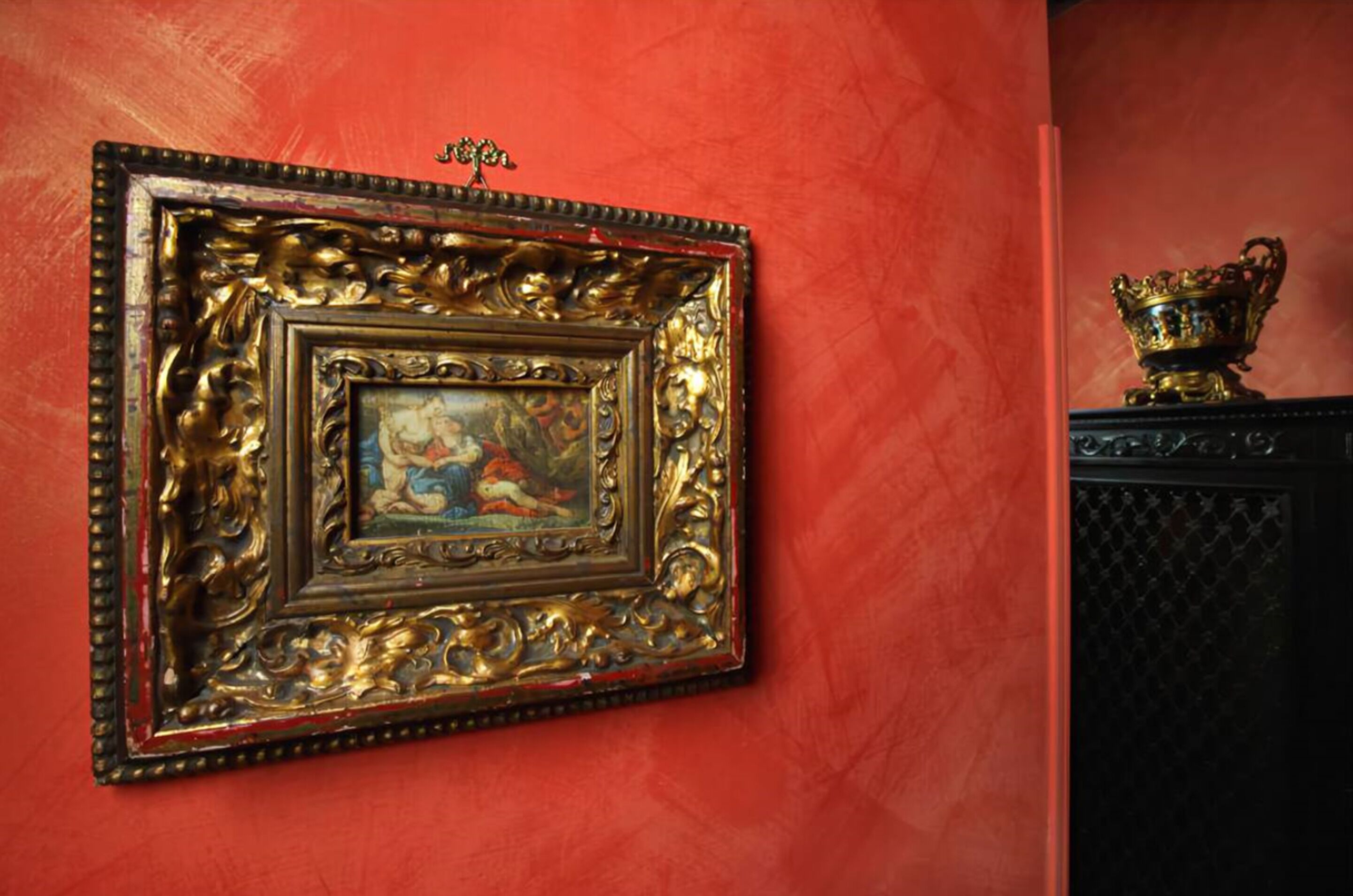 Una obra de arte enmarcada sobre una pared roja, un objeto dorado decorativo sobre una superficie negra y un pequeño adorno dorado encima de la obra.