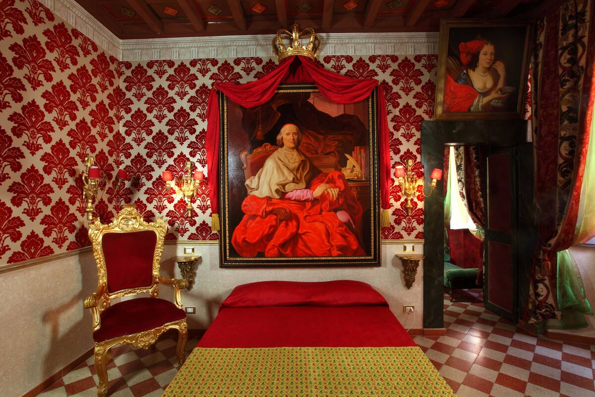 A room with a large portrait, a red bedspread, a gold chair, and patterned wallpaper.