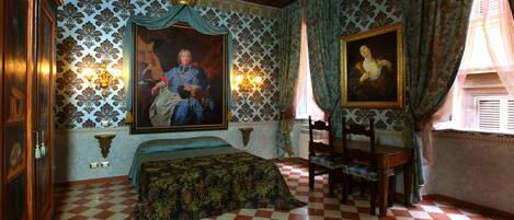 A room with a checkered floor, a bed with a patterned bedspread, a wooden chair, a wooden table, and two wall-mounted lamps.