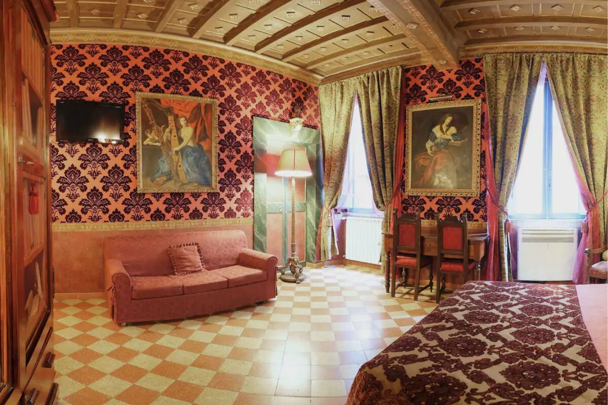 A room with a checkered floor, a pink sofa, a bed with a patterned bedspread, a table with chairs, and two paintings on the walls.