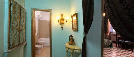 A narrow hallway with a checkered floor, a bathroom with a toilet and sink, and a mirror with wall sconces.