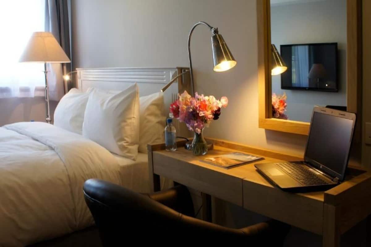 Habitación de hotel con una cama, un escritorio con una computadora portátil, una lámpara, un florero con flores y un televisor en la pared.