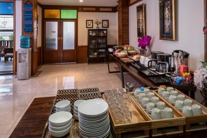 A hotel lobby with a reception desk, a water dispenser, a coffee station, and a buffet area with plates and glasses.