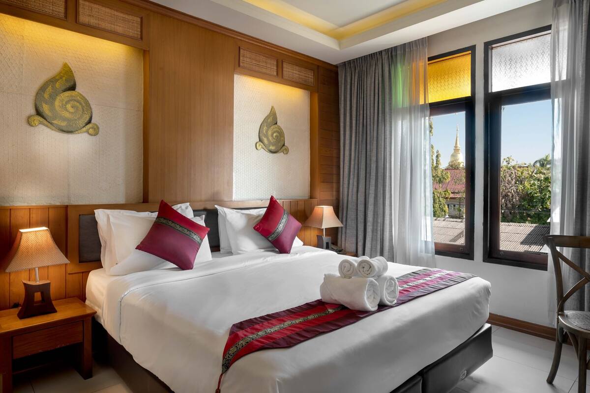 A hotel room with a large bed, two bedside lamps, a view of a temple through the window, and a wooden headboard.