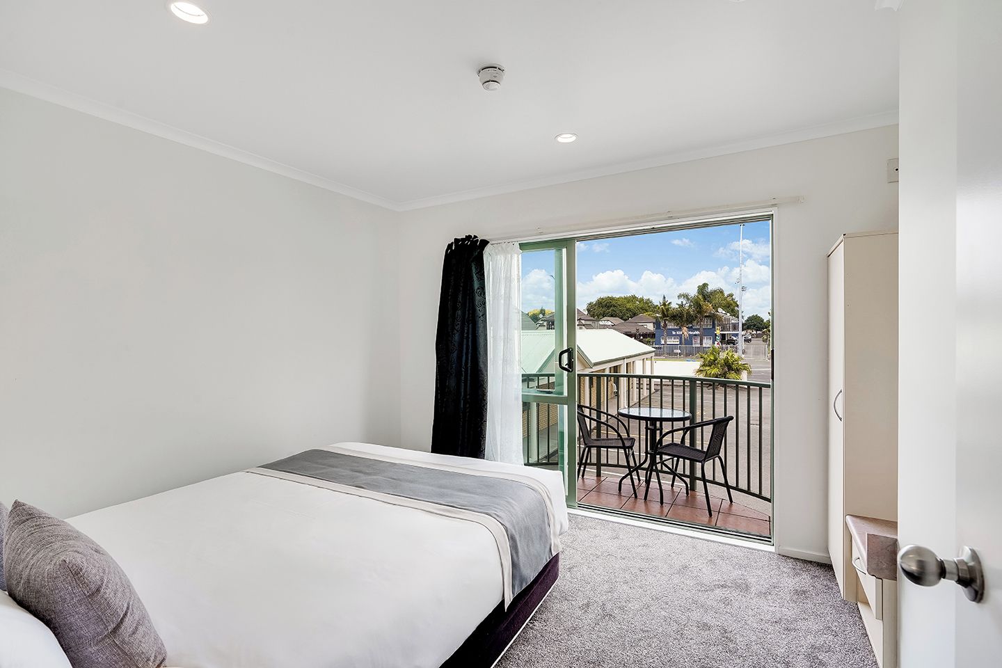 Quarto de hotel com cama, varanda com mesa e cadeiras, e vista para a rua.