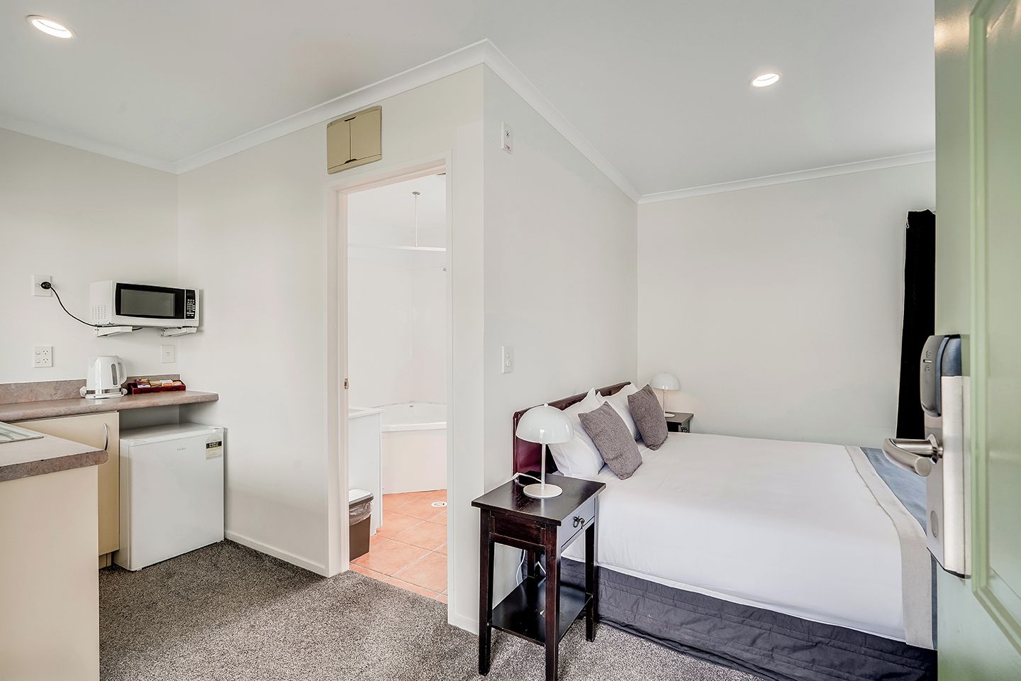 Habitación de hotel pequeña con kitchenette, una cama con almohadas, una mesita de noche y una puerta que da a un baño.