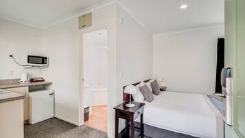 Habitación de hotel pequeña con kitchenette, una cama con almohadas, una mesita de noche y una puerta que da a un baño.
