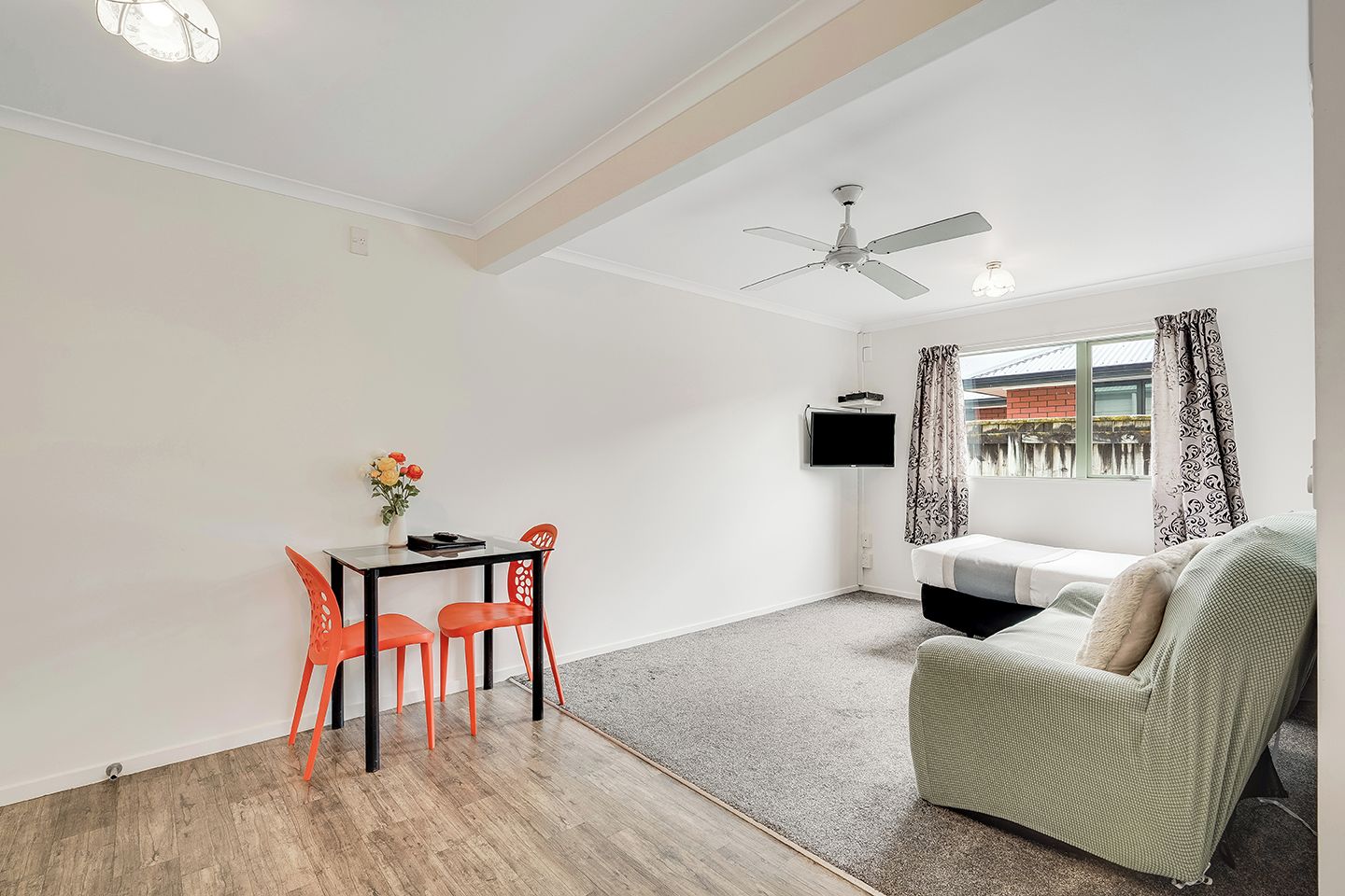 A room with a dining table, chairs, a sofa, a ceiling fan, a TV, and a window with curtains.