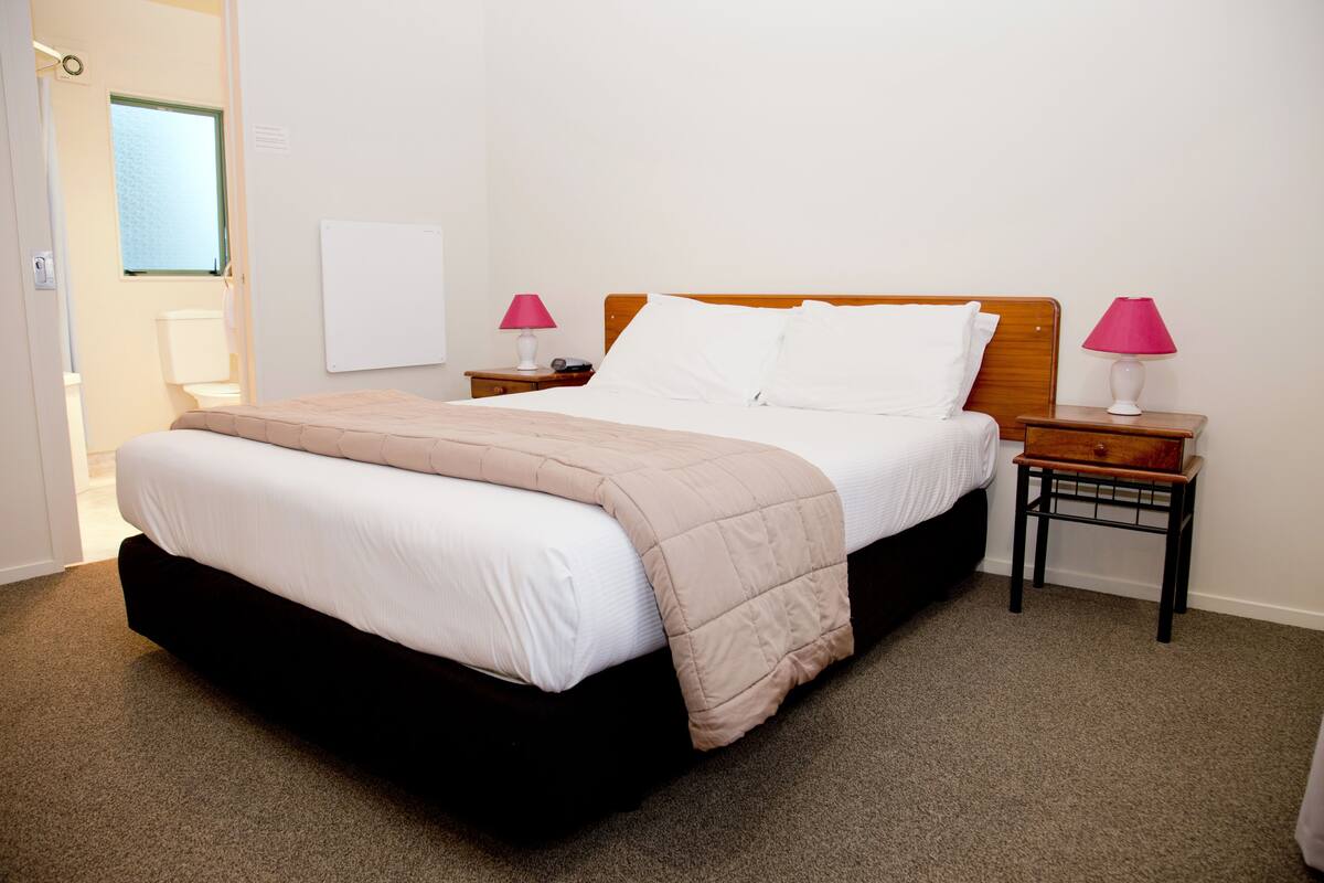 A neatly made bed with a beige blanket, two bedside tables with lamps, a whiteboard on the wall, and a window with blinds.