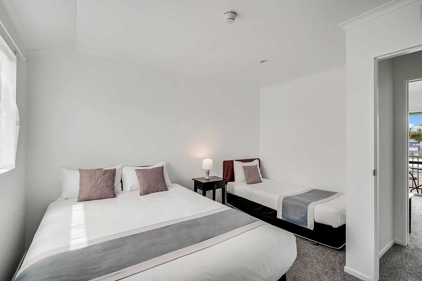 Quarto de hotel com uma cama grande, uma cama menor, mesa de cabeceira com luminária e vista para o exterior.