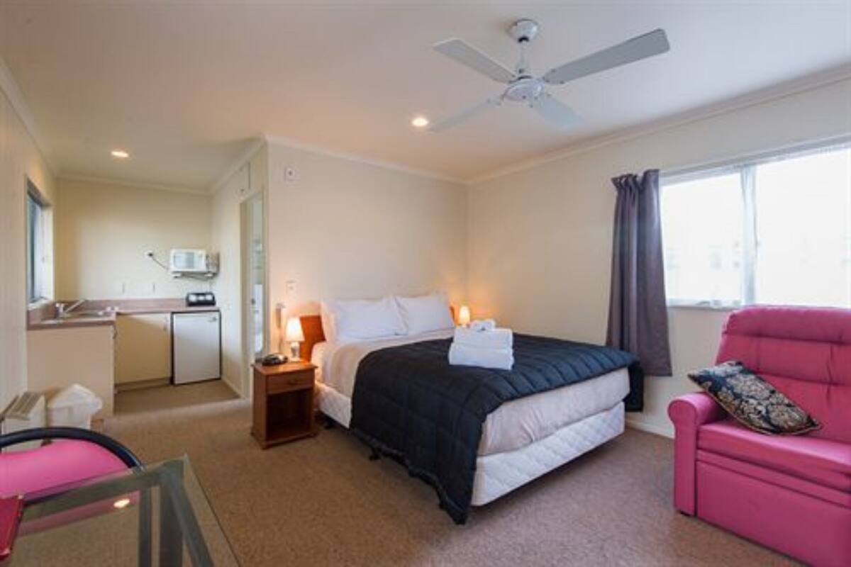A compact hotel room with a single bed, a small desk with a chair, a kitchenette, and a window with curtains.
