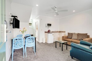A compact living space with a kitchenette, a dining area with blue chairs, a sofa with patterned cushions, and a ceiling fan. - Hamilton Settlers Motor Lodge (Hamilton)
