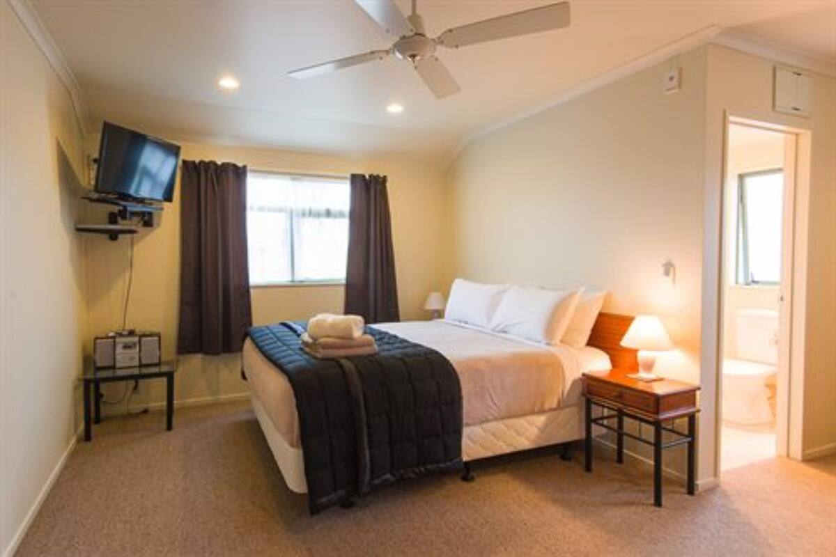 A bedroom with a bed, a ceiling fan, a TV mounted on the wall, a bedside table with a lamp, and a window with curtains.