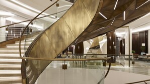 Un escalier en colimaçon moderne, doté de rampes en verre et d’une finition métallique, s’élève au centre d’un vaste atrium à plafond haut, éclairé par un système d’éclairage contemporain.