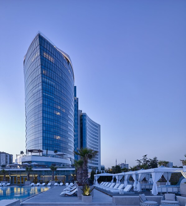 A modern hotel with a curved glass facade, a rooftop pool with sun loungers, and a white canopy structure.