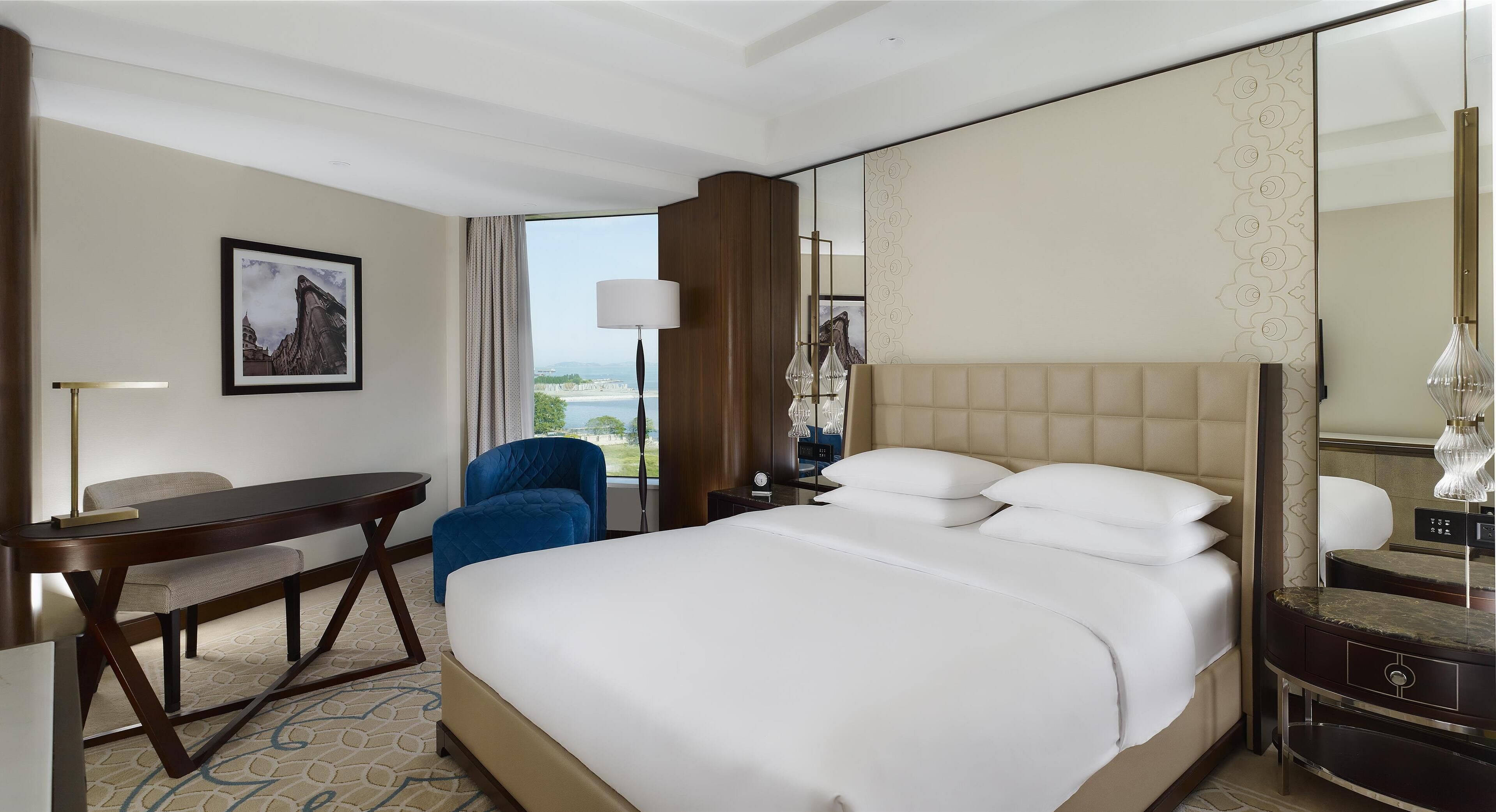 A hotel room with a bed, a desk, a chair, a lamp, and a large window with a view.
