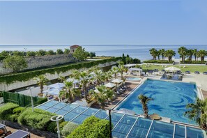 A rooftop pool with clear blue water, surrounded by palm trees and lounge chairs, overlooking a calm sea and a stone wall.