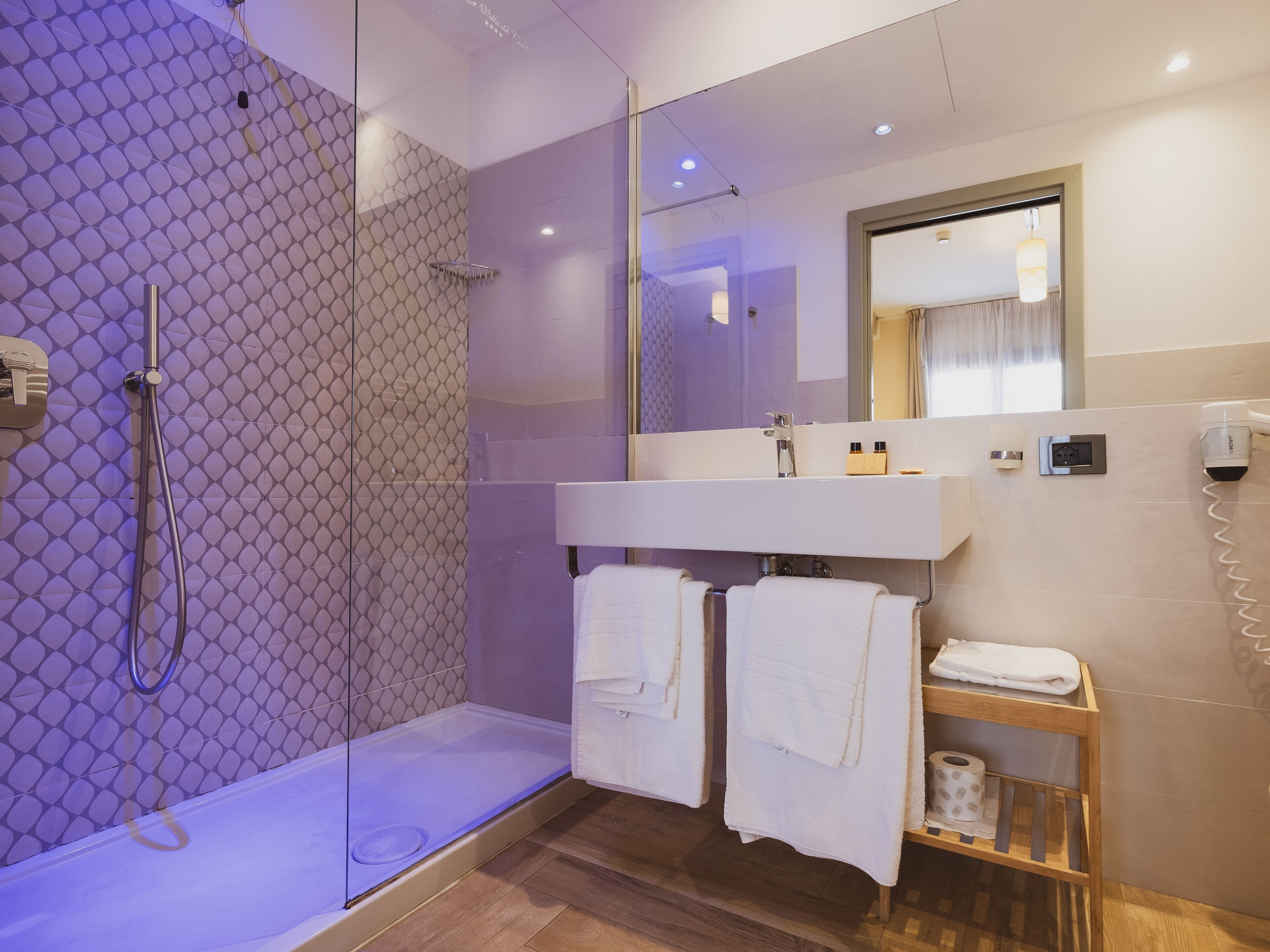 Une salle de bain moderne avec une douche à parois de verre, un meuble-lavabo avec un miroir et une étagère en bois où sont rangées des serviettes et des articles de toilette.
