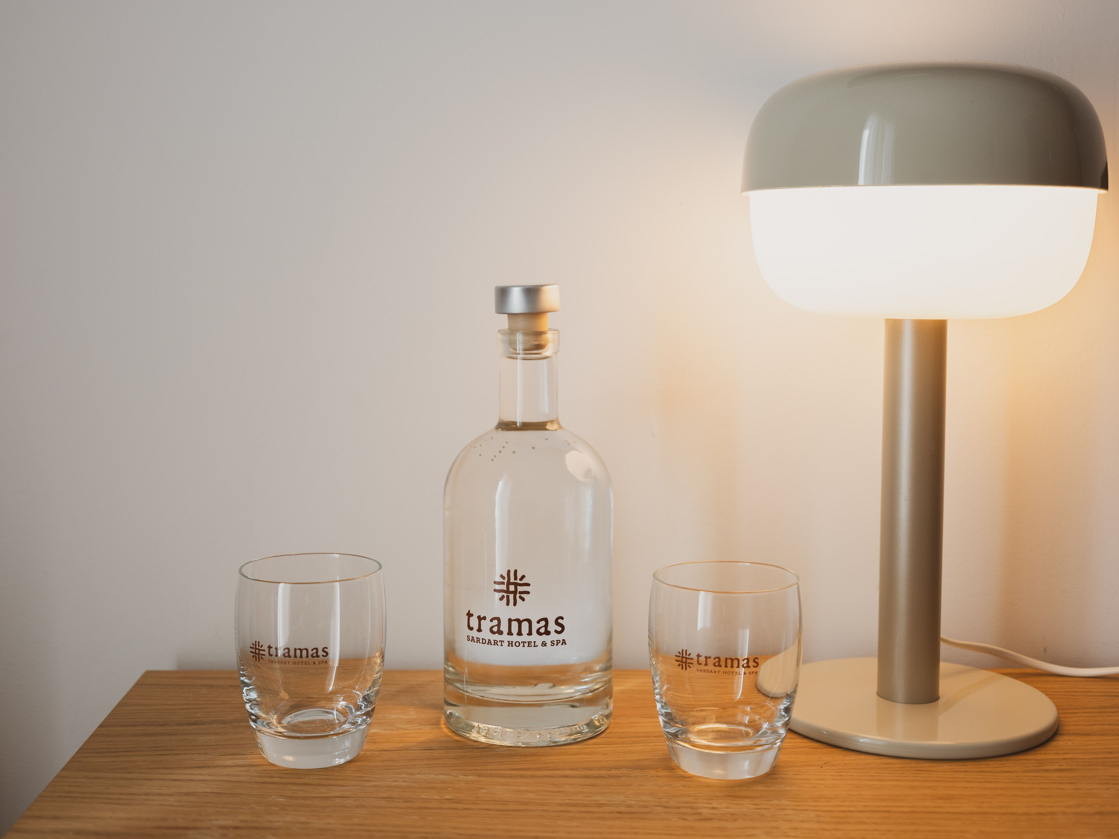 A bottle of Tramas Sardiart Hotel & Spa, two glasses, and a lamp on a wooden table.