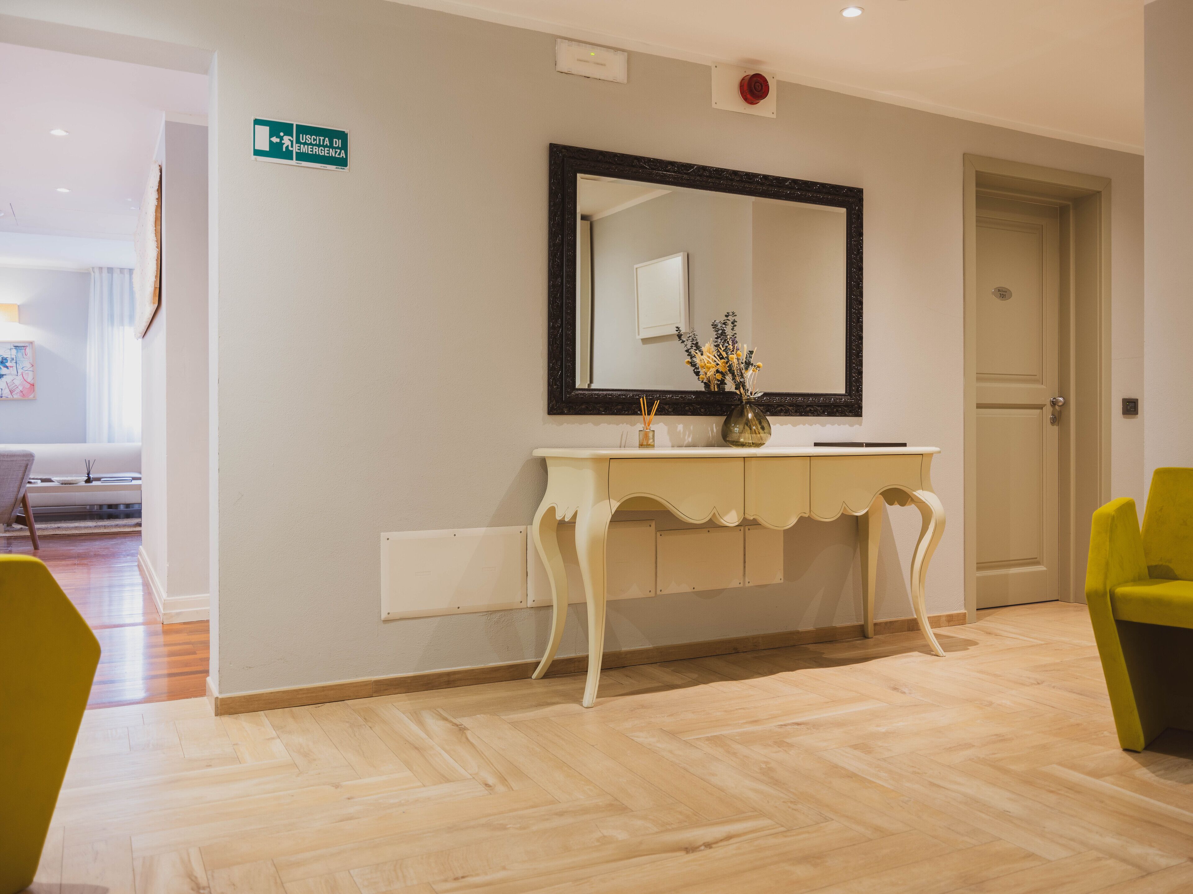 A hotel reception area with a white console table, a large mirror, and a vase with flowers. There is a door to the right and a hallway leading to another room.