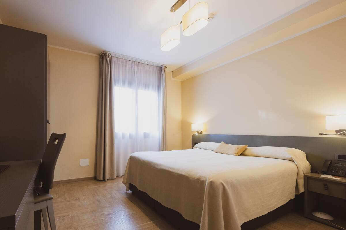 A hotel room with a bed, bedside table, lamp, and a desk with a chair.