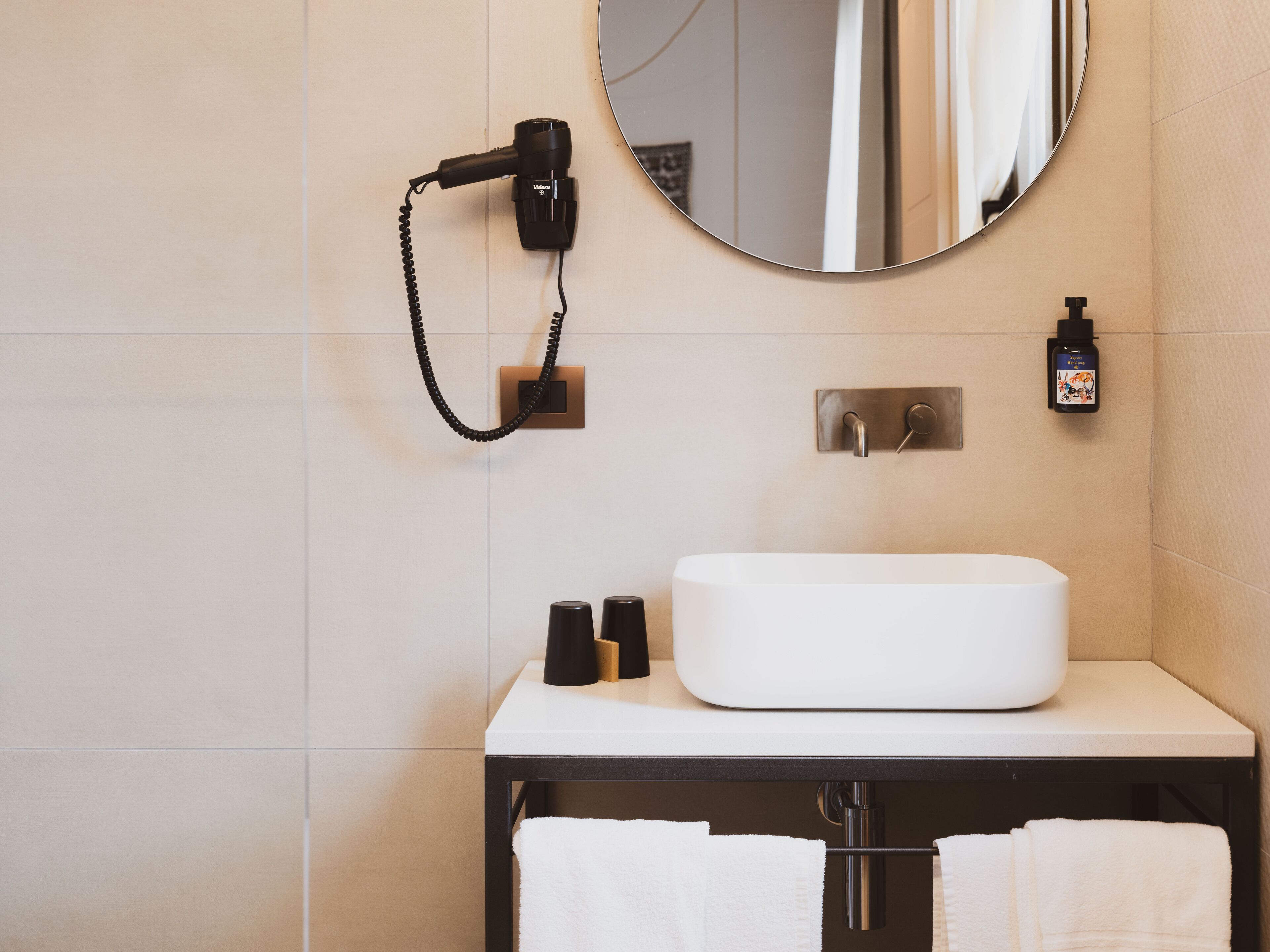Un baño moderno con un lavamanos blanco, un espejo redondo, un secador de pelo y un dispensador de jabón para manos.