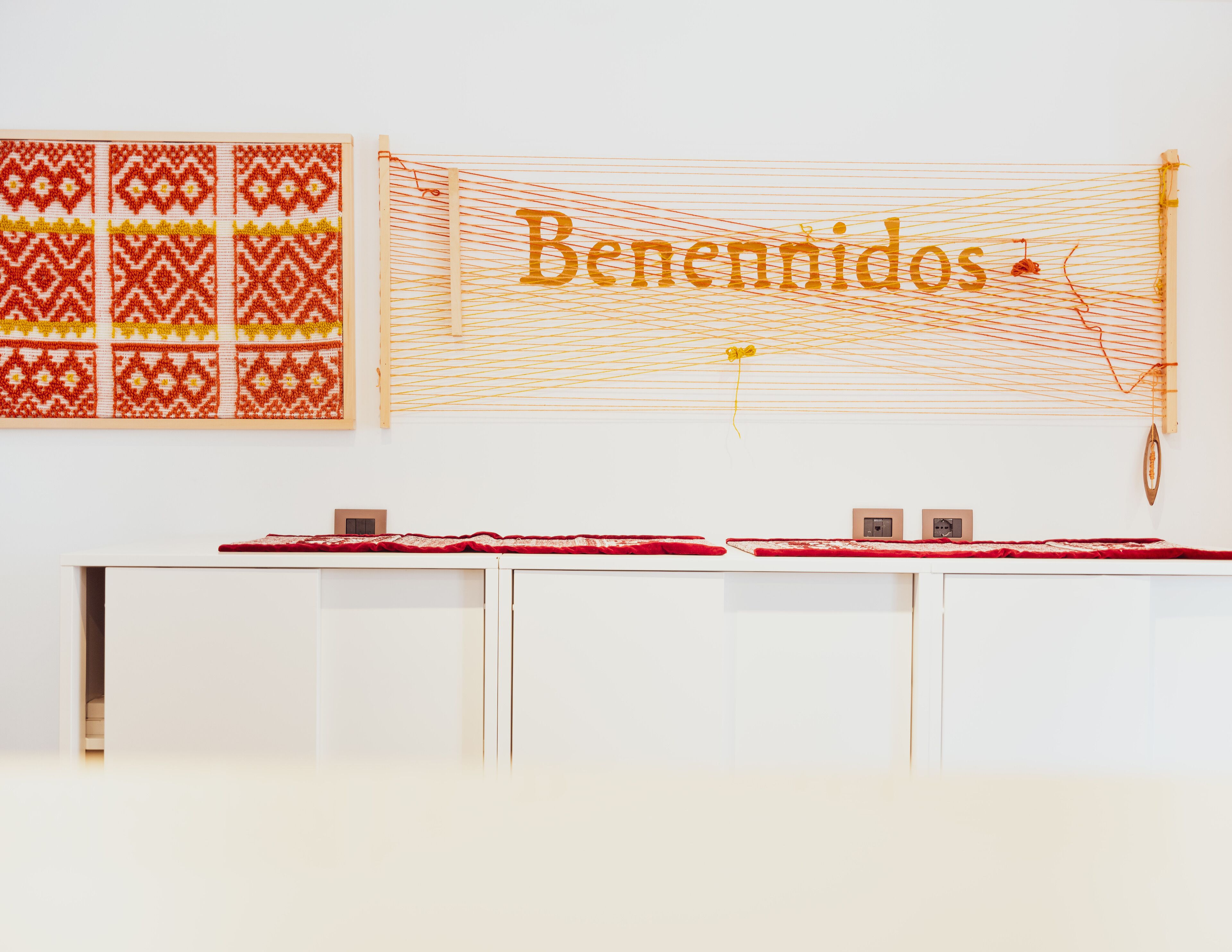 A room with a white wall, a patterned textile on the left, and a large 'Benennidos' sign on the right.