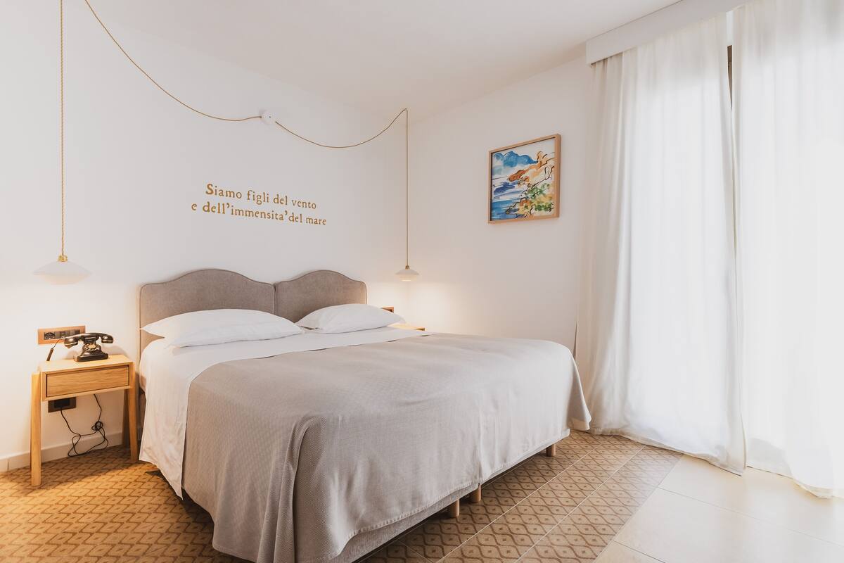 A bedroom with a bed, bedside tables, a wall-mounted artwork, and a window with curtains.