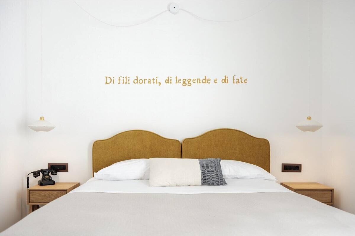A neatly made bed with a wooden headboard, white bedding, and a decorative pillow. There are two bedside tables with lamps and a wall-mounted light fixture.