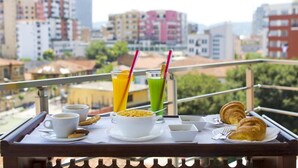 Ein Frühstückstablett mit Kaffee, Croissants und zwei Getränken auf einem Balkon mit Blick auf eine Stadtlandschaft.