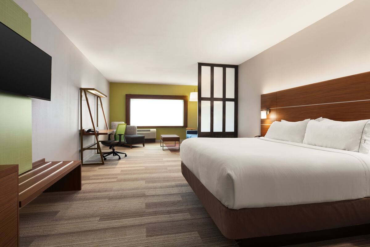 A hotel room with a large bed, a desk with a chair, a television, and a window with blinds.