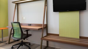 Un espacio de oficina moderno con un escritorio de pie, una silla de oficina ergonómica, un televisor de pantalla plana montado en la pared y una pequeña mesa redonda.