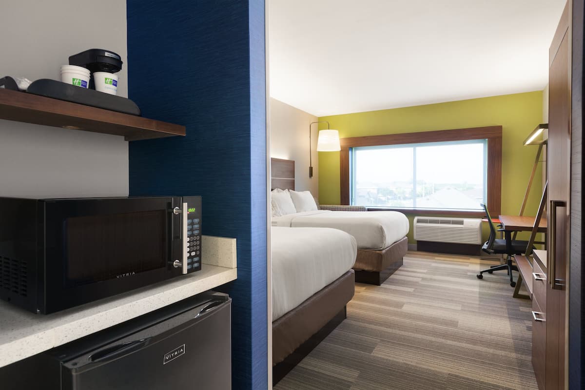 A hotel room with a microwave, mini-fridge, two beds, a desk, and a large window.
