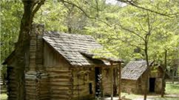 A rustic log cabin with a porch and a smaller structure nearby, surrounded by trees and greenery. - Holiday Inn Express & Suites Tahlequah by IHG (Tahlequah)