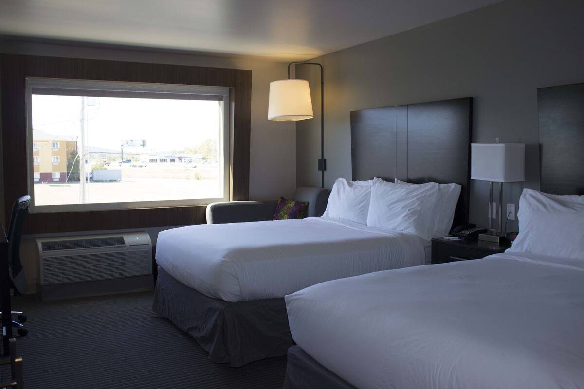 A hotel room with two beds, a desk, a chair, a lamp, and a window with a view of buildings outside.