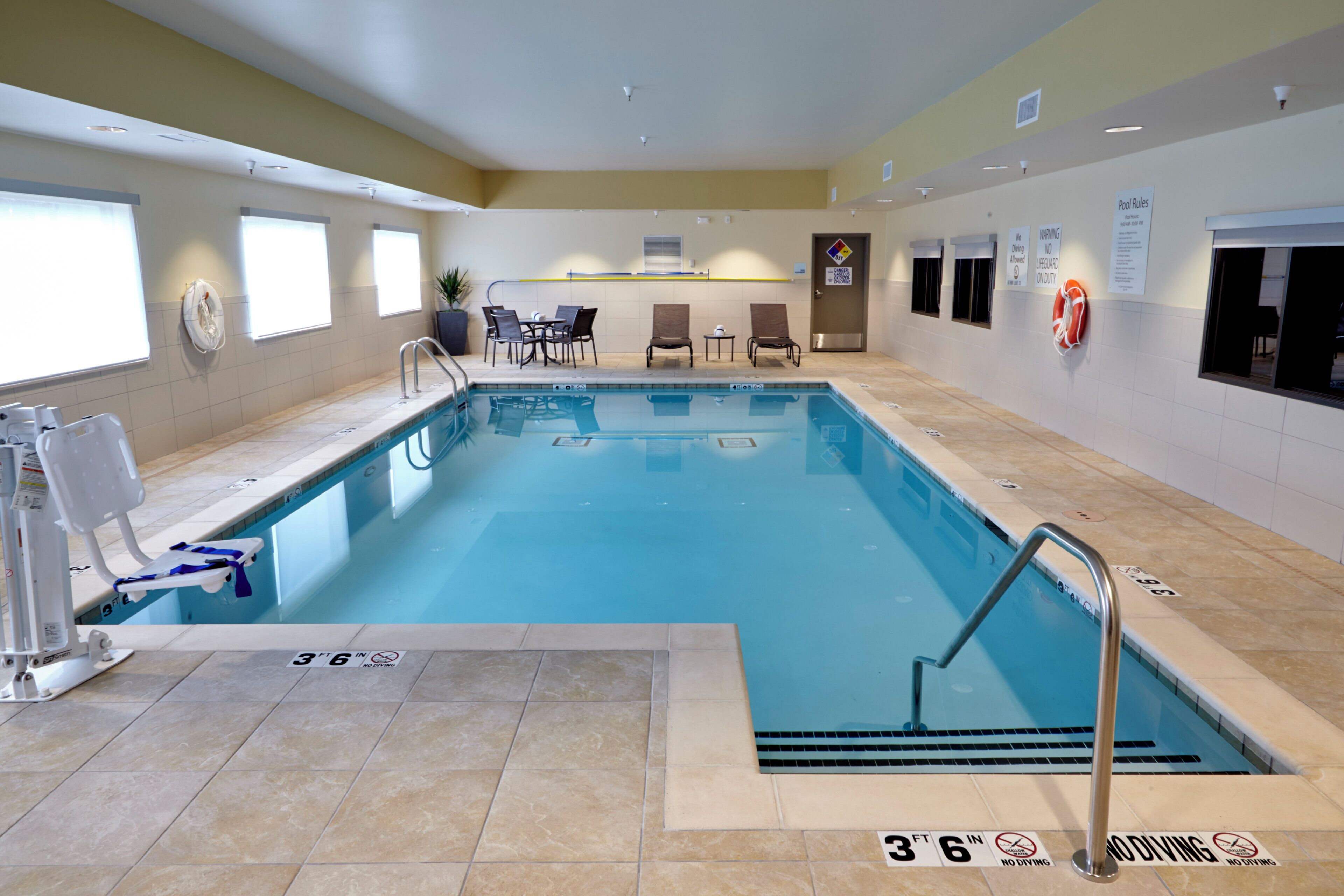 Una piscina interior con un asiento de socorrista, carteles con las normas de la piscina y una zona de estar con sillas y mesas.