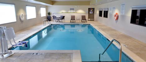 Una piscina interior con un asiento de socorrista, carteles con las normas de la piscina y una zona de estar con sillas y mesas.