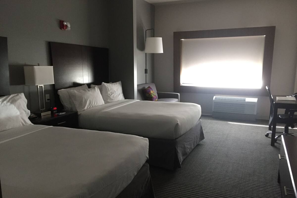 A hotel room with two beds, a desk, a chair, a lamp, and a large window.