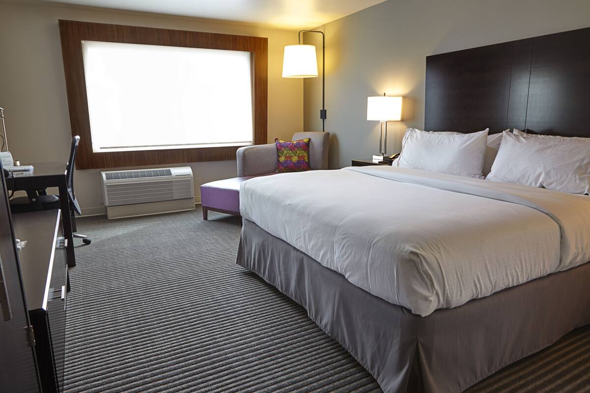A hotel room with a large bed, a desk, a chair, a window, and a wall-mounted air conditioner.