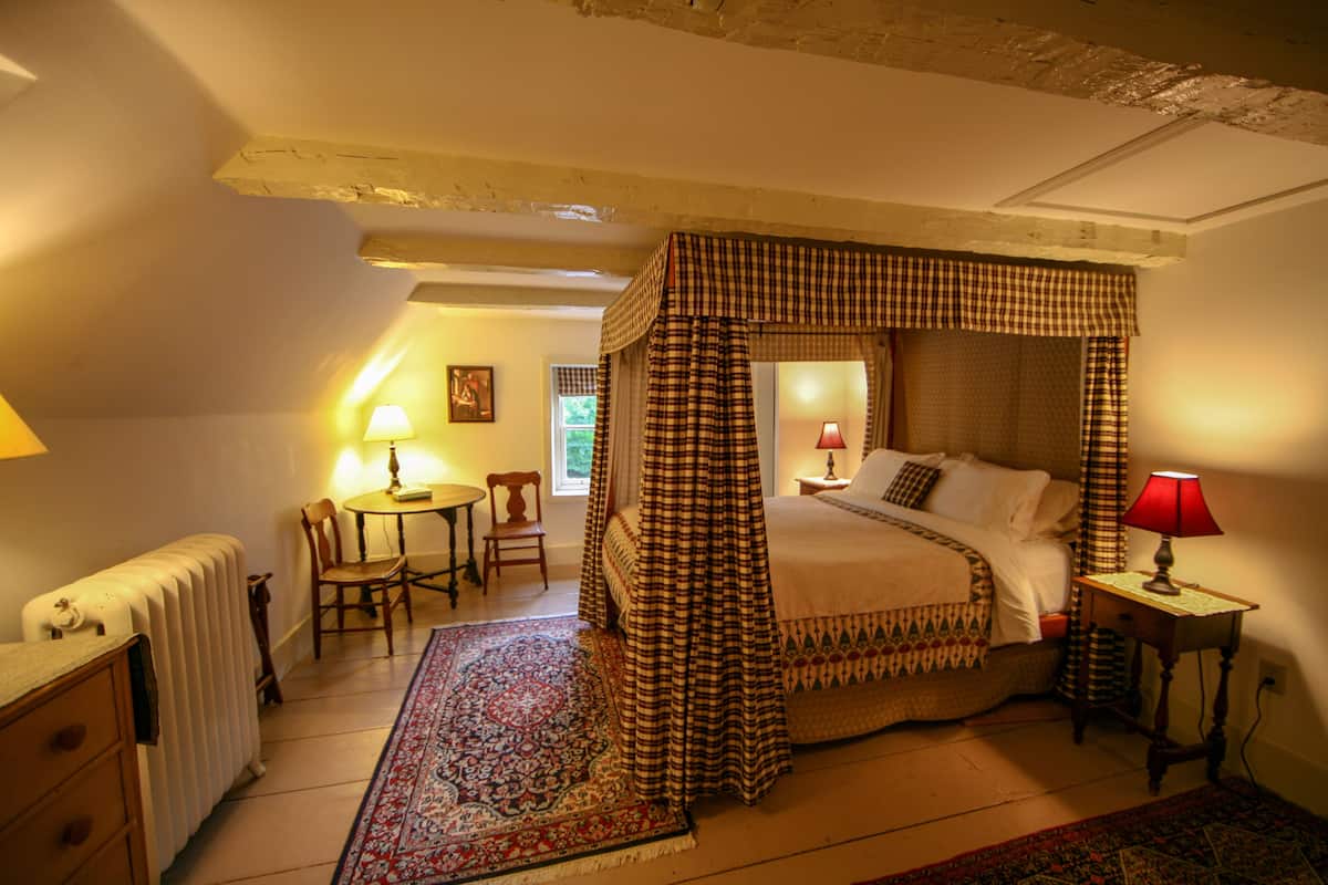 A four-poster canopy bed with a checkered bedspread, a red lamp on a bedside table, a wooden chair, a small round table with a lamp, and a radiator.