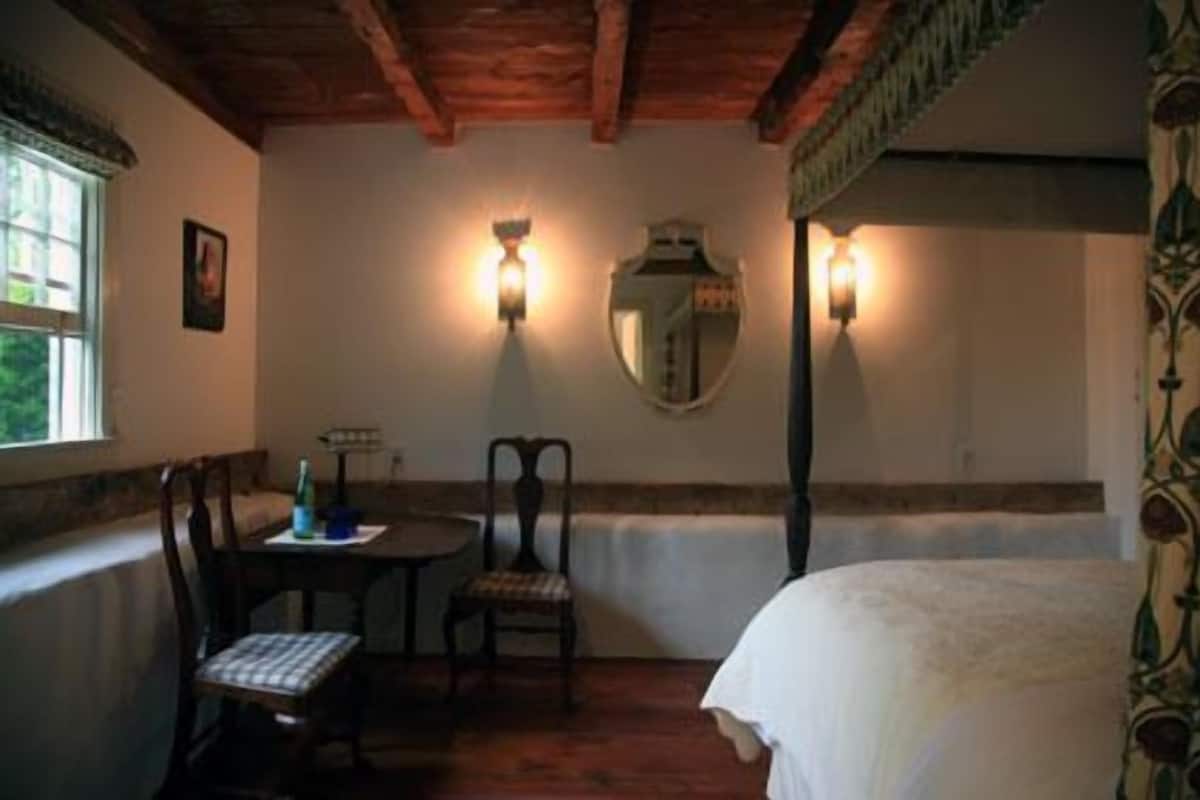 A bedroom with a wooden ceiling, a bed, a table with chairs, and two wall-mounted lights.