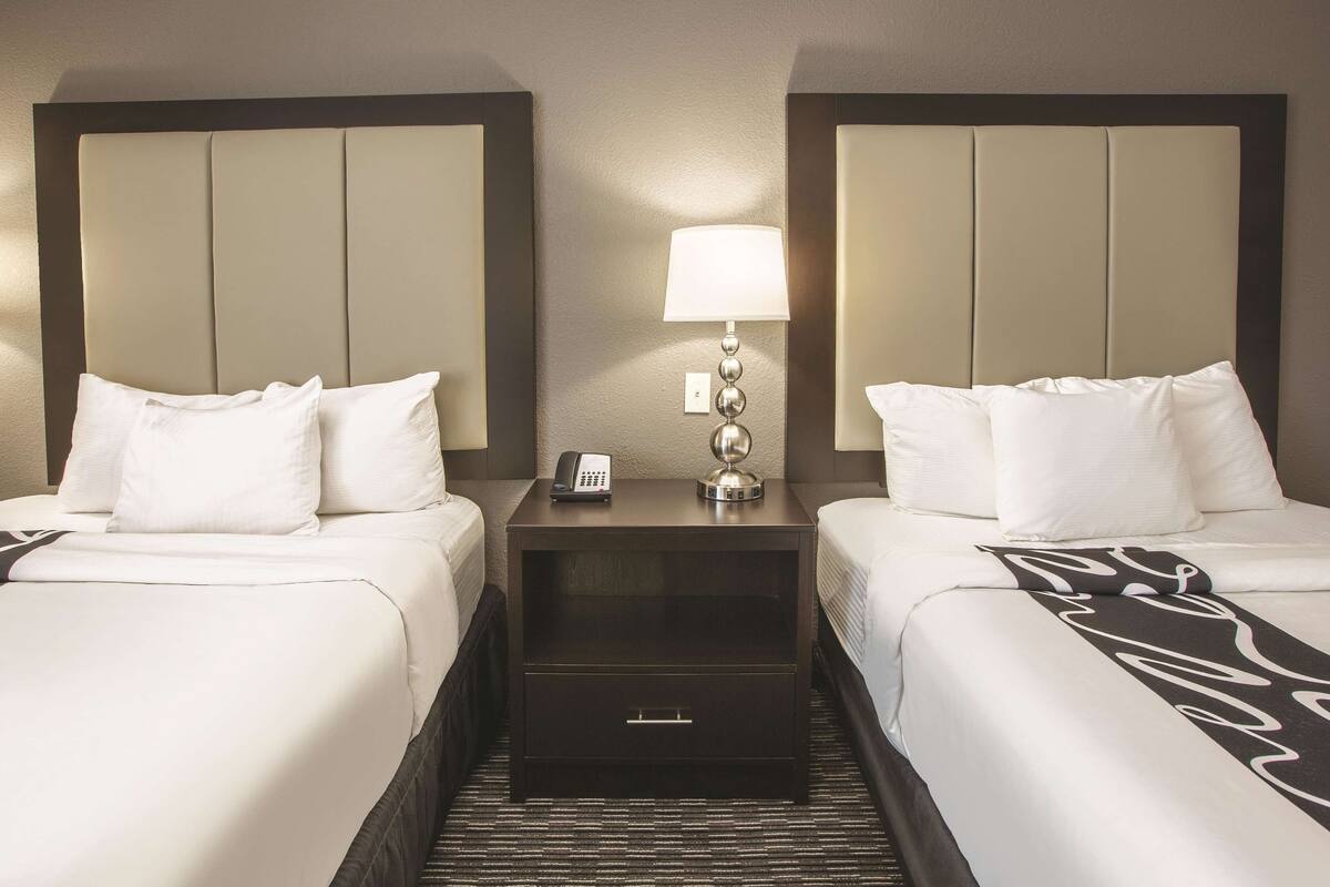 A hotel room with two beds, a nightstand with a lamp, a phone, and a drawer.