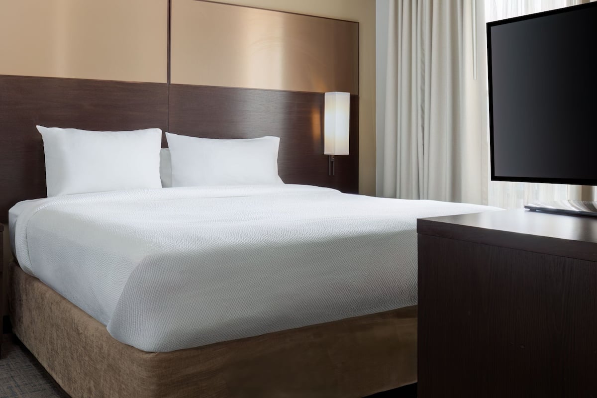 A neatly made bed with white linens and a wooden headboard, a bedside lamp, and a flat-screen TV on a wooden dresser.